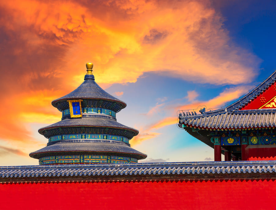 El Templo del Cielo en Pekín detrás de un muro rojo tradicional, bajo un cielo brillante con nubes de color naranja y amarillo durante el atardecer.